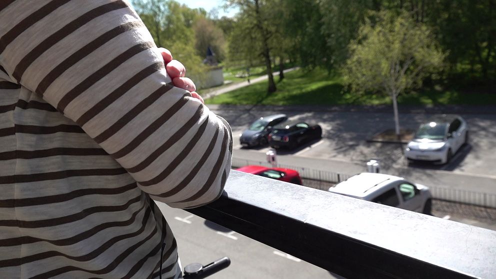 Person i randig tröja lutar sig mot balkongräcke och tittar ner mot parkering och grönområde.