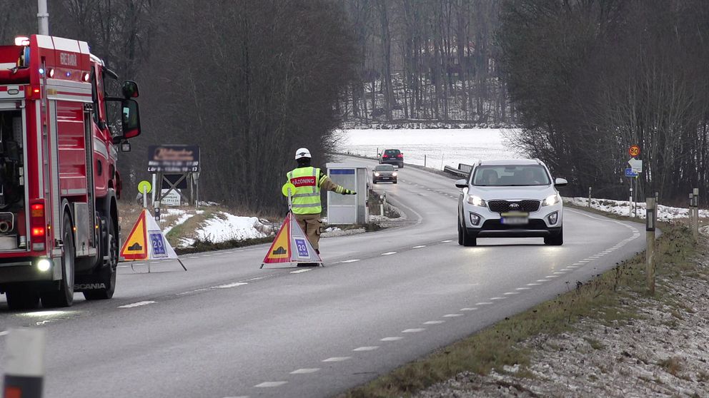 Väg 204, på vägen står en räddningstjänstbil och skyltar med texten olycka. En brandman gör tecken till trafiken att sänka hastigheten.