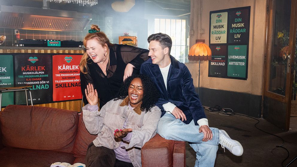 Linnea Wikblad, Emil Hansius och Assia Dahir sitter på en soffa i en informell miljö med affischer för Musikhjälpen i bakgrunden. Amie gestikulerar med handen, medan de andra två poserar avslappnat.