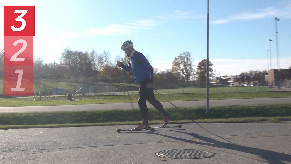 En person åker längdskidor på en asfaltväg under klar himmel. I vänster kant finns en nedräkning som visar siffrorna 3, 2, 1. I bakgrunden kan man se grönområden och sportanläggningar.