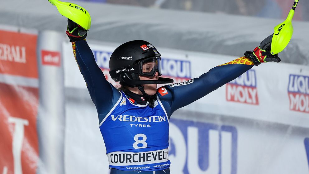En alpin skidåkare i tävlingsdräkt firar med armarna i luften i Courchevel med startnummer 8. Reklambanderoller syns i bakgrunden.