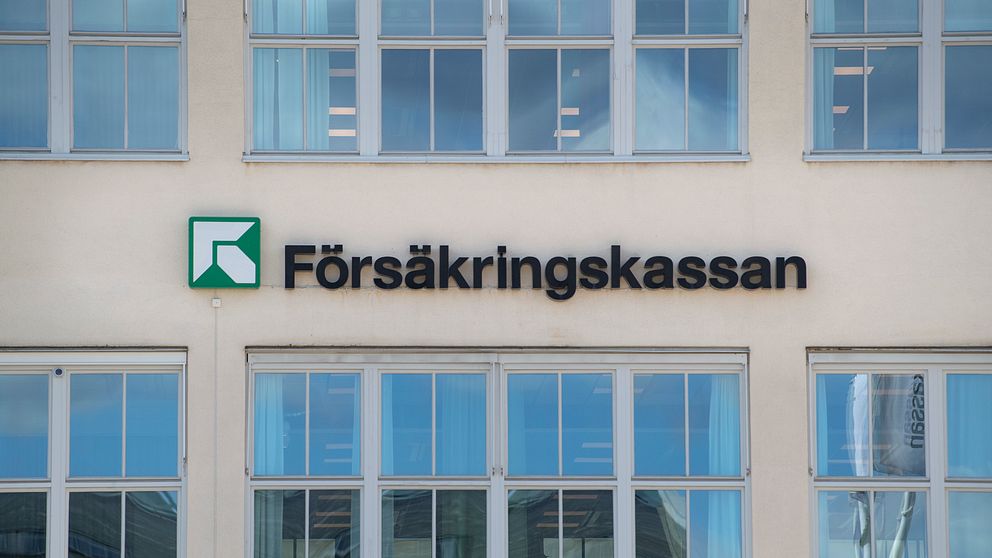 Skylt på en byggnad med texten ”Försäkringskassan” och en grön logotyp. Flera fönster syns i bakgrunden, vilket ger en modern kontorsmiljö.