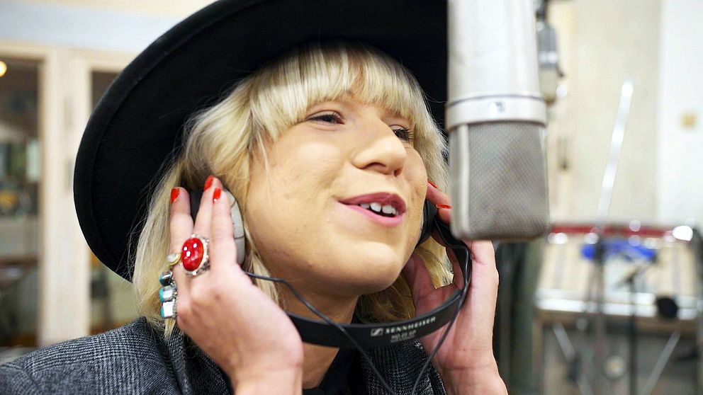 Ellinor står med ljust hår, svart hatt, röda naglar och en stor röd ring vid en mikrofon och håller hörlurar vid öronen