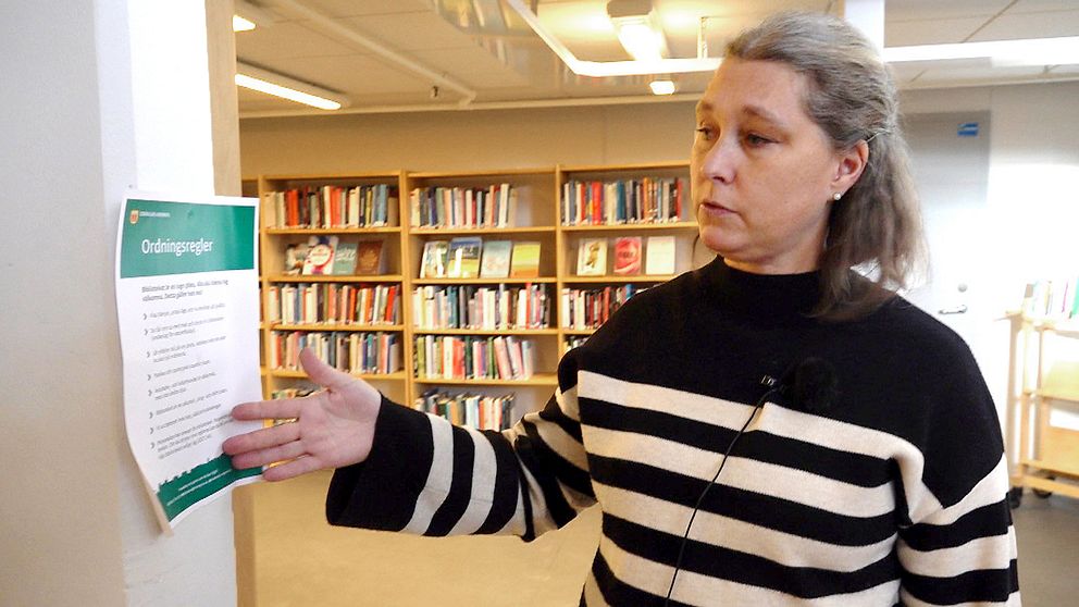 Bibliotekschefen Towe Karlsson står i biblioteket och pekar på en ordningsskylt