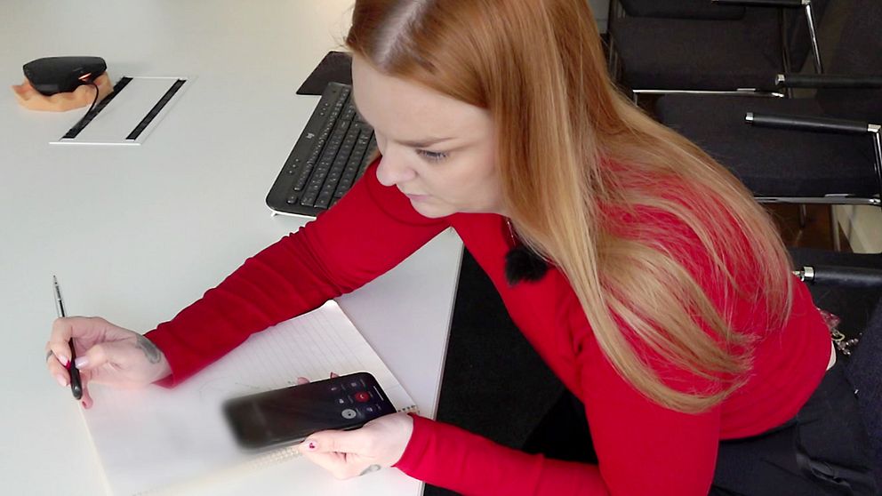SVT:s reporter Anna Wallbrandt sitter med pappaer och penna och pratar i telefon med högtalare på. Hon har en röd tröja på sig och rödlätt hår.