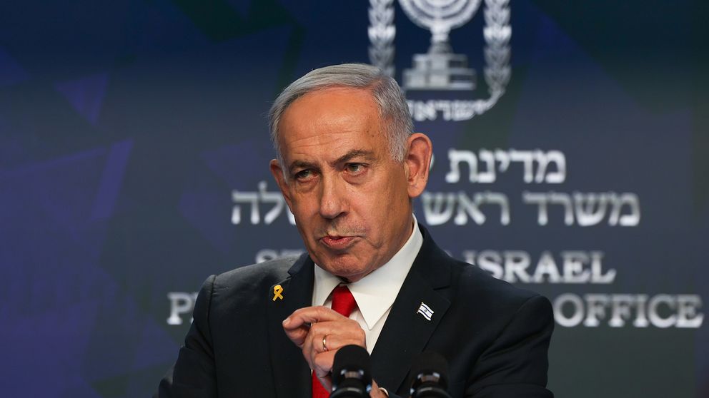 En person i kostym talar vid ett podium med Israels statsvapen i bakgrunden.