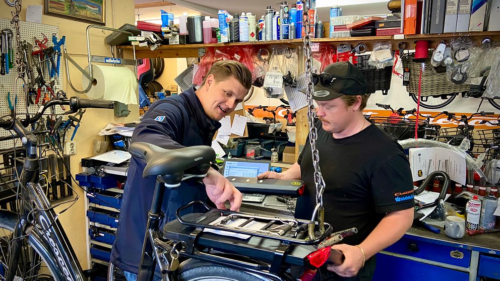 Två personer samarbetar vid reparation av en elcykel i en verkstad fylld med verktyg.