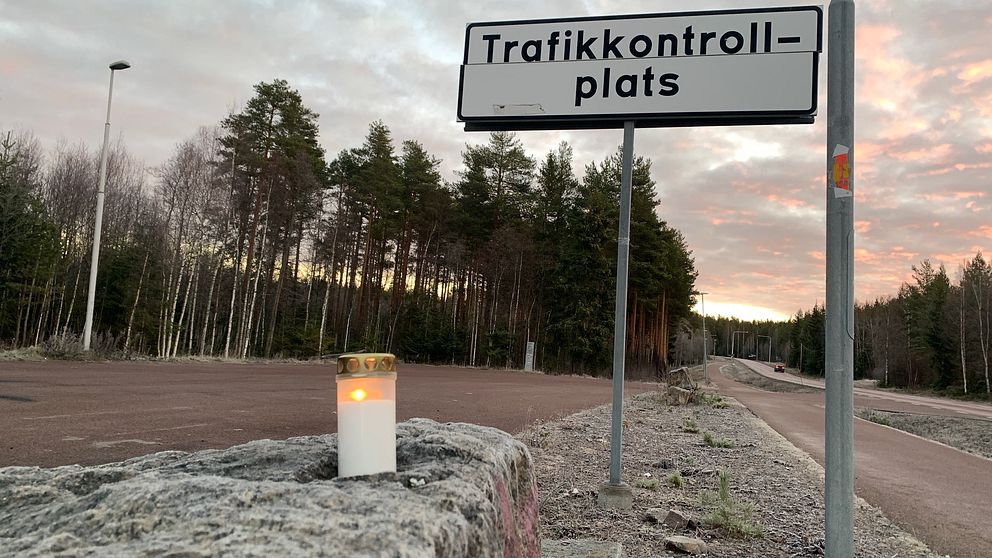 En vägskylt som visar ”Trafikkontrollplats” står nära en parkeringsyta omgiven av träd, med en tänd ljuslykta placerad på en sten i förgrunden. Skyn är färgad.