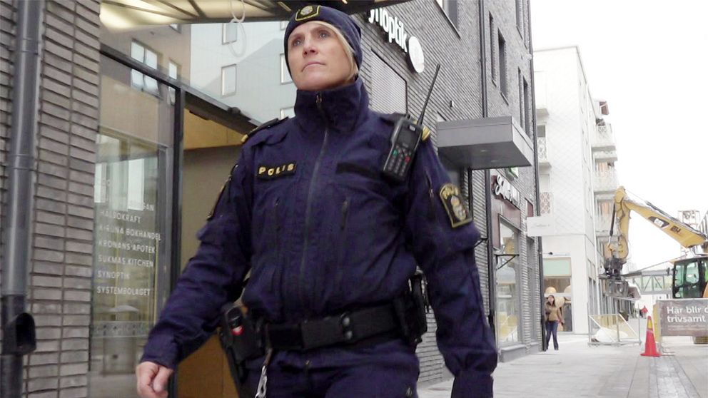 En polis i uniform, Therese Forslund, går längs en gata i Kiruna centrum. Flera byggnader syns i bakgrunden. I nära anslutning finns en entré till en butik, och längre bort syns en traktor gräva i en gata.