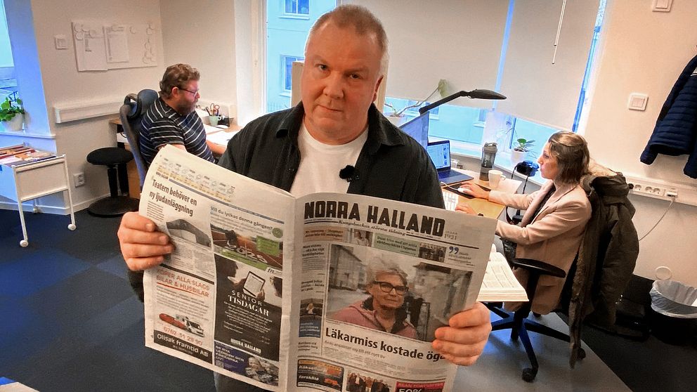 En person håller upp en tidning med titeln ”Norra Halland” i ett kontorsutrymme där två andra personer arbetar vid skrivbord. Tidningsrubrikerna är synliga, och rummet har naturligt ljus som strömmar in från ett fönster.