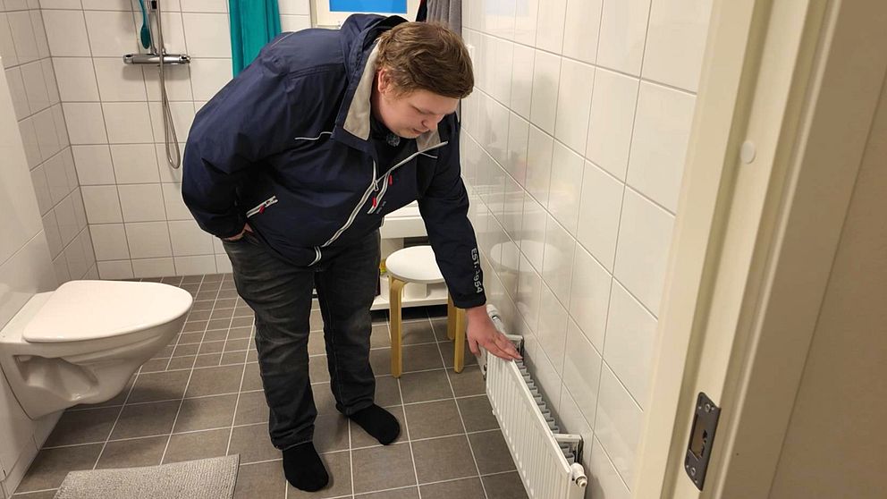 Jonatan Ståhl står i sitt badrum och pekar på en radiator längs väggen. I bakgrunden syns en toalett och en stol. Badrummet har ljusa väggar och en grå keramikplattläggning på golvet.