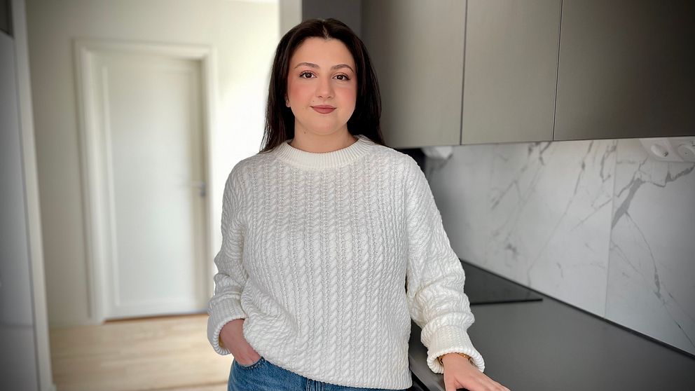 Person i vit stickad tröja står i ett modernt kök med gråa skåp och marmormönstrad vägg.