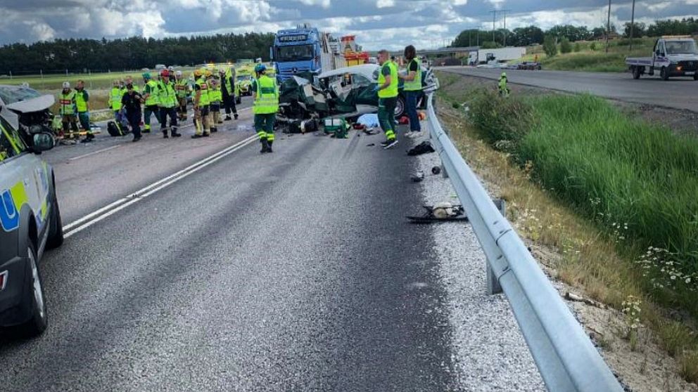 En trafikolycka på en motorväg med flera räddningspersonal som arbetar vid olycksplatsen. Två bilar är involverade, varav en verkar ha skadats allvarligt. Räddningstjänst och ambulanspersonal är synliga, och vägen är avspärrad. I bakgrunden syns trafik.