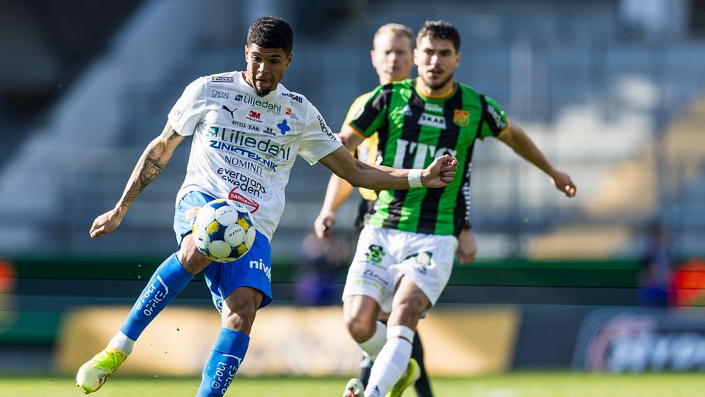 Fotbollsspelare i vit och blå dräkt sparkar bollen, motståndare i grönsvart förföljer.