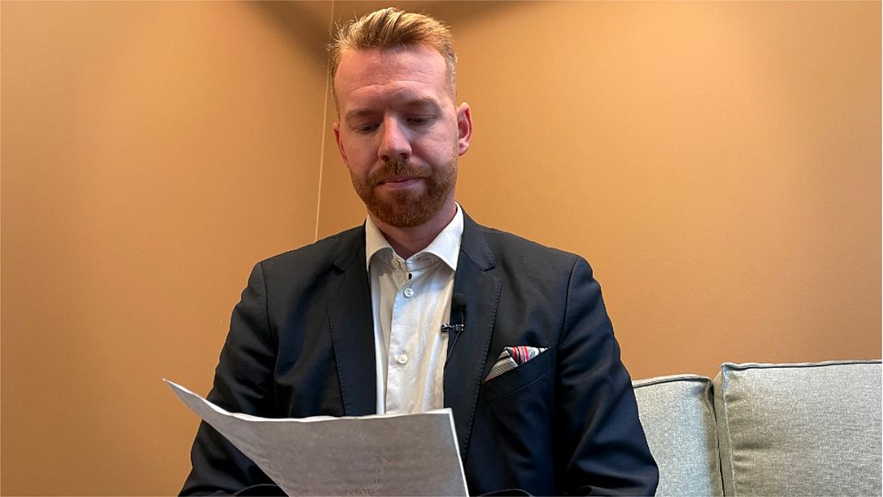 En man i kostym sitter i en soffa och håller i ett papper. Han är fotograferad från en frontvinkel i en inomhusmiljö med väggar i en varm nyans. Det finns en kudde i bakgrunden.