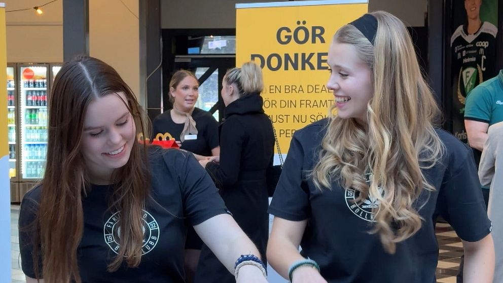 Två personer i svarta tröjor står vid ett bord fyllt med små kartonger eller burkar i en köpcentrummiljö. Bakom dem står skyltar med texten ”Gör Donken!” och information om jobbansökningar hos McDonald's.