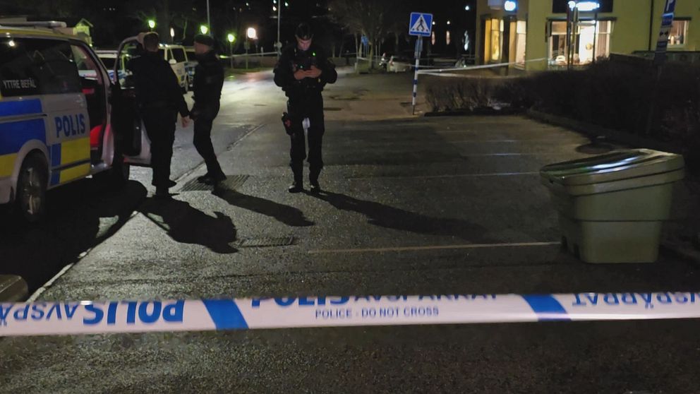 Poliser står i en upplyst parkeringsplats där en polisbil är parkerad. En av poliserna använder en mobiltelefon medan de andra samtalar. En avspärrning med texten ”POLIS – DO NOT CROSS” syns i förgrunden. I bakgrunden finns en sopkärl och en affärslokal i svagt ljus.