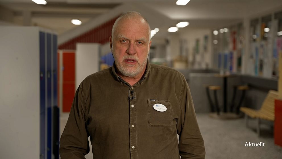 En person i brunt skjorta står framför en skola med blå och röda skåp i bakgrunden, vilket skapar en miljö som antyder en utbildningsinrättning.