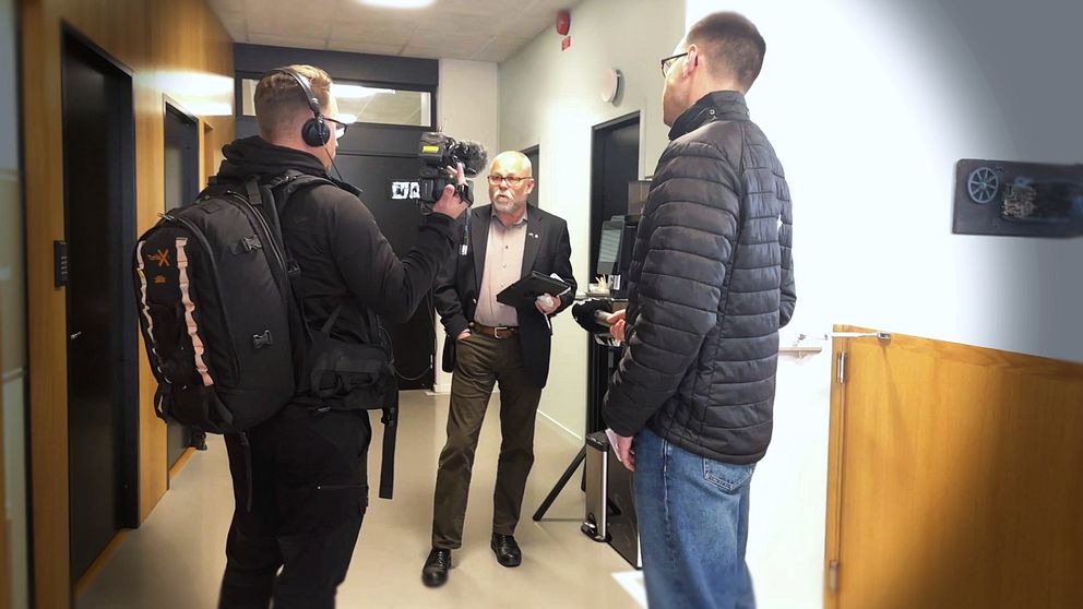 En reporter och en kamerateam står i en korridor, där reportern intervjuar en person som håller i papper. Den tredje personen observerar från sidan. Korridoren har ljusa väggar och dörrar på vardera sidan.