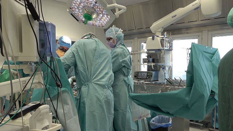 Kirurgisk operation som pågår i ett operationsrum. Flera sjukvårdspersonal i skyddsutrustning arbetar tillsammans kring en patient på operationsbordet. Utrustning och monitors syns i bakgrunden, och det råder en koncentrerad atmosfär.