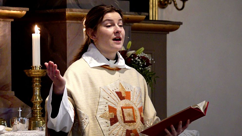En präst, 24-åriga Evelina Sjöwall, klädd i en ornamenterad mässhake håller en öppen bok och verkar vara mitt i en predikan i en kyrka. En tänd ljusstake syns i bakgrunden.