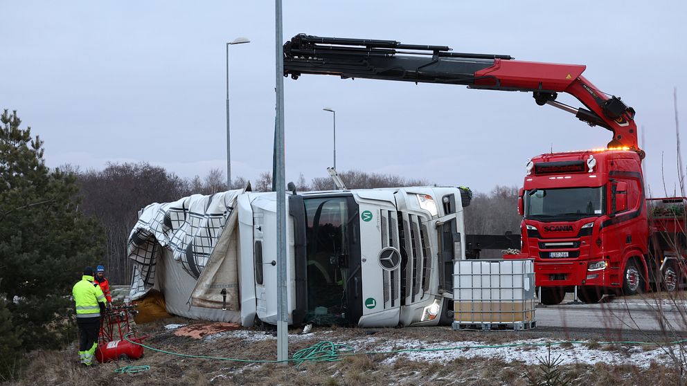 En vält lastbil ligger på sidan vid vägkanten. En kranbil är på plats för att assistera med uppröjningen, och en person i varselkläder syns arbeta nära olycksplatsen.