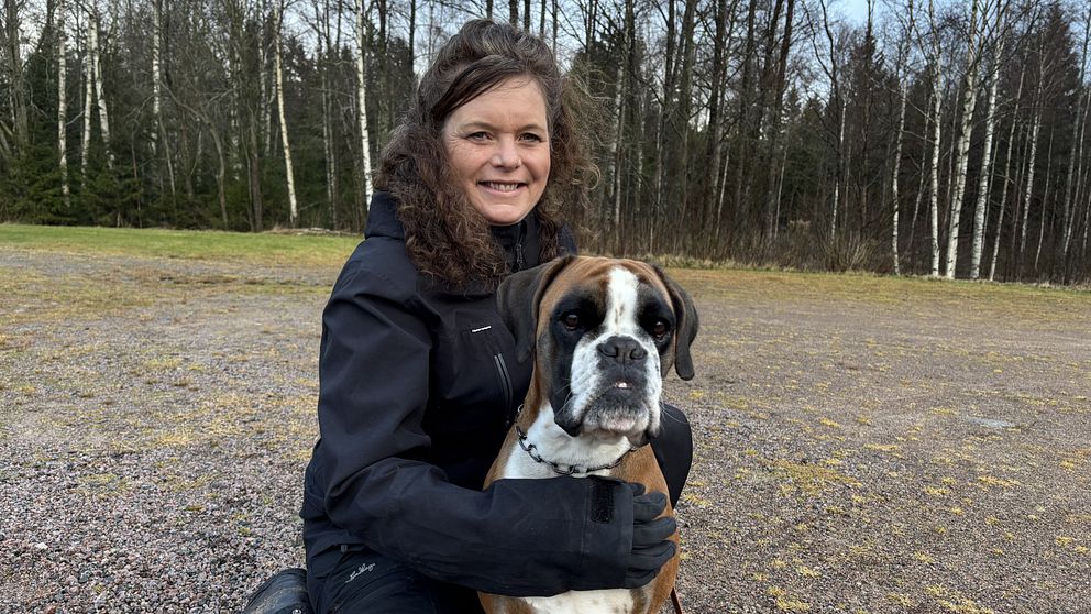 Veronica Frohlund sitter på marken med en boxerhund, omgiven av en skogsliknande miljö med träd i bakgrunden. Personen bär en svart jacka och kramar om hunden med ena armen. Hundens uttryck är vänligt och nyfiket.