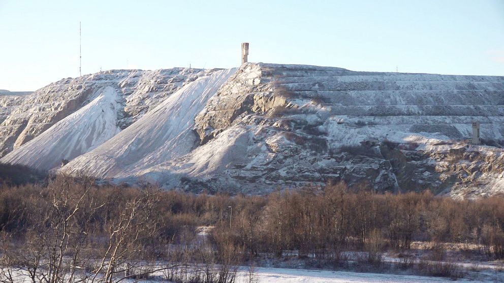En snötäckt bergsformation med branta sidor, toppad av ett tornliknande struktur, omgiven av ett landskap med snö och trädkronor.