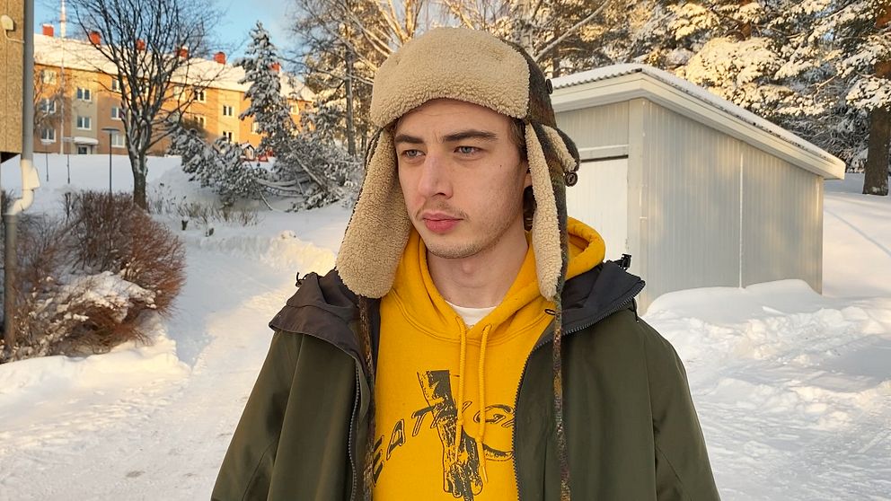 En person står i en snöig miljö iförd en gul sweatshirt och en varm mössa med öronlappar. Bakgrunden består av snötäckta träd och flerfamiljshus som syns i avståndet.