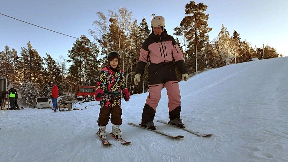 Ett barn och en vuxen står i en skidbacke, bägge har hjälm. Barnet har en blommig overall, mamman en skiddräkt i svart och rosa.