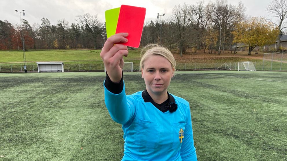 Kvinna i blå domartröja står på fotbollsplan och visar upp ett gult och ett rött kort.