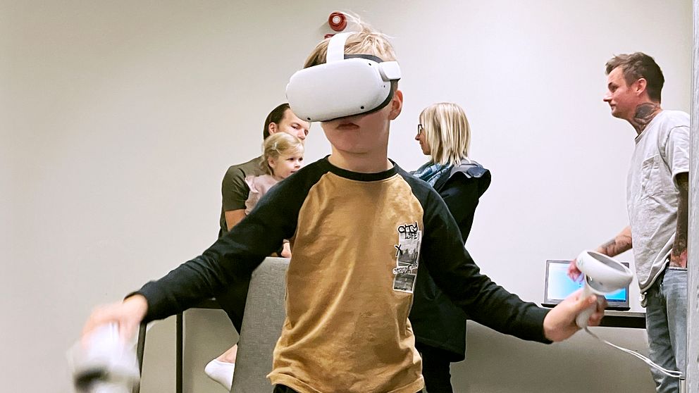 En pojke med VR-headset utforskar en virtuell verklighet medan han står i ett rum. I bakgrunden finns flera personer som observerar honom och ett skrivbord med en dator.