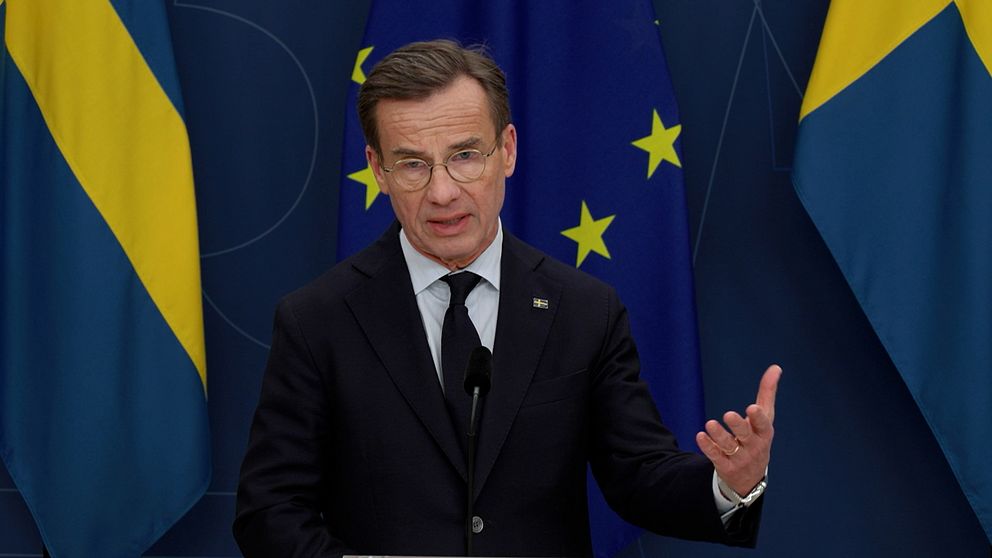 Ulf Kristersson (M) står och pratar vid ett podium framför en blå bakgrund med Sverige- och EU-flaggor i Regeringskansliets pressrum.