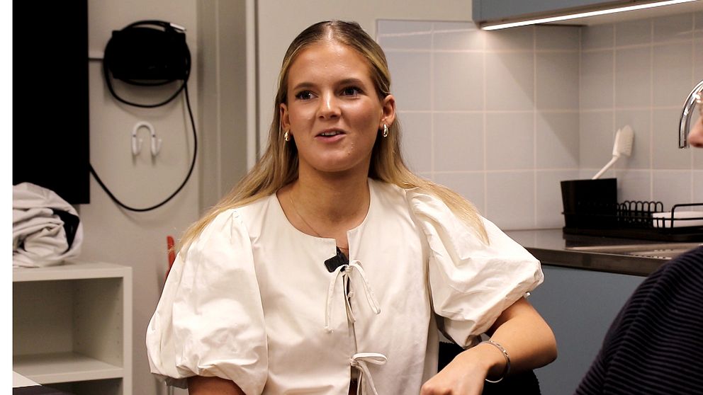 En kvinna med långt hår sitter i en inomhusmiljö i en ljus blus.