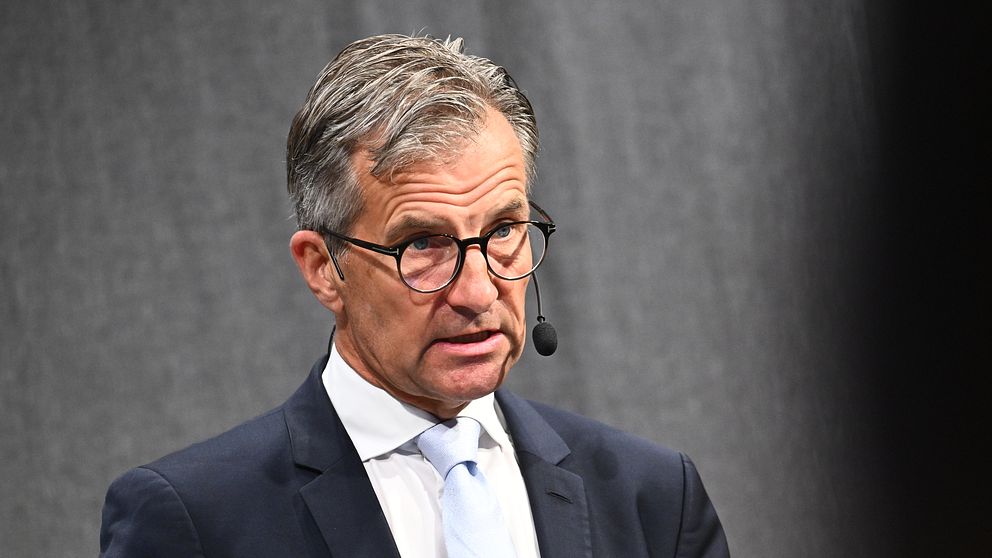 Erik Thedéen står på scenen och talar i en mikrofon. Han bär en mörk kostym med en vit skjorta och en ljusblå slips. Bakgrunden är en grå scen.