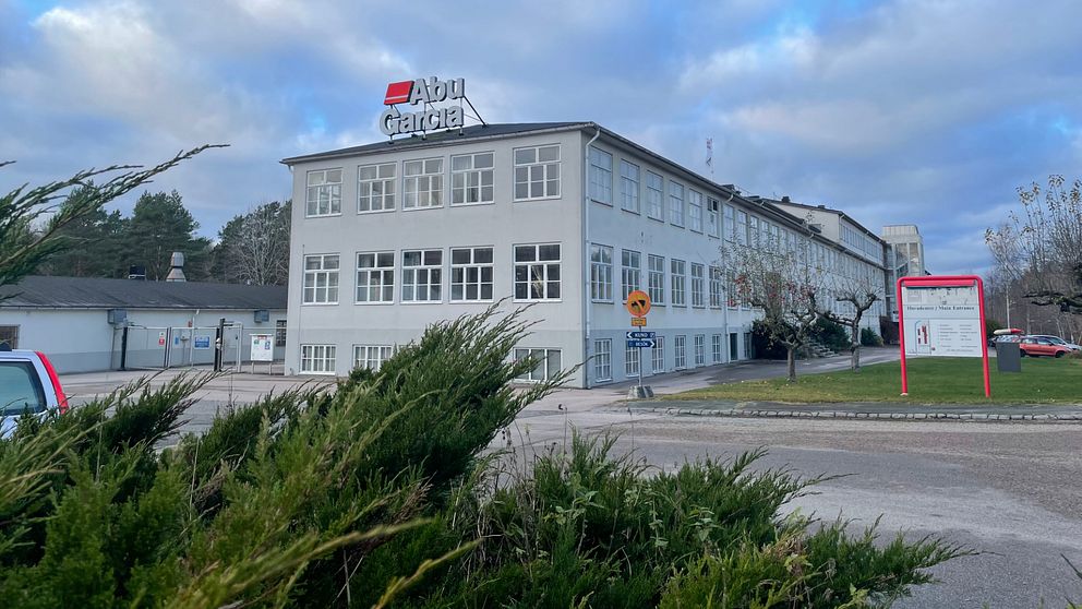 Byggnaden med skylten ”Abu Garcia” i tydliga bokstäver, omgiven av buskar och träd. Det är en grå, fler våningsbyggnad med stora fönster. Himlen är molnig och vädret verkar kallt. En vägskylt och en väg kan ses i förgrunden, vilket indikerar att bilden är tagen nära en väg.
