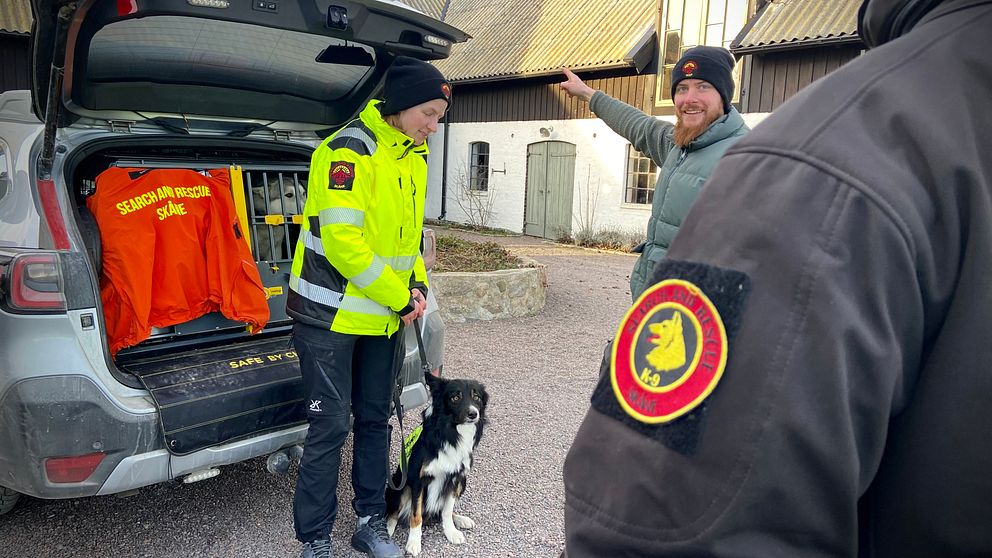 En räddningstjänstmedlem i en gul reflexjacka står vid en öppen bil med hunden kopplad vid sin sida, medan en annan person gestikulerar med handen. I bilen syns orangea tröjor med text som indikerar räddningsverksamhet.