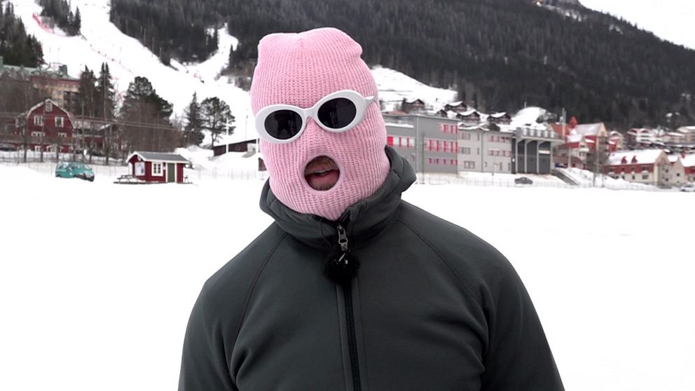 Person i mörk vinterjacka och rosa mask står utomhus på en snöklädd plats i Åre. Åre memes.