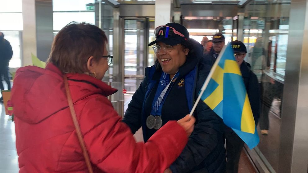 Person i röd jacka överlämnar svensk flagga till medaljprydd individ i vintermiljö.