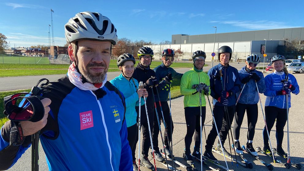 En grupp av åtta skidåkare i färgglada träningskläder står på en asfalterad yta, hållandes skidor och stavar. De är samlade i en rad med en klar blå himmel och en industriell byggnad i bakgrunden. Stämningen verkar vara avslappnad och vänlig, vilket antyder en gemensam träning eller aktivitet.