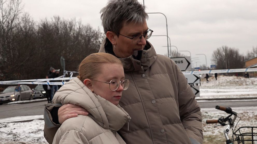 Två personer står utomhus iförda tjocka vinterjackor. Området bakom dem är avspärrat med polistejp och det finns några polisbilar synliga i bakgrunden. Det är en kall vinterdag med snö på marken.