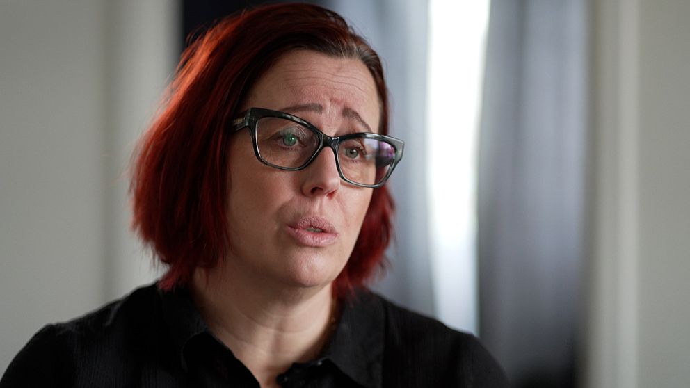 En person med rödbrunt hår iklädd i en svart skjorta, stående inomhus med ett dunkelt belyst bakgrund.