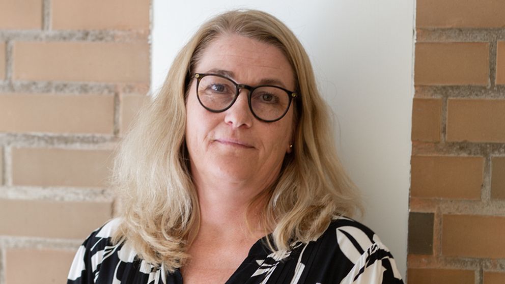 Kvinnlig person med blont, vågigt hår, klädd i en svart och vit blommig blus, står framför en ljus vägg av sten.