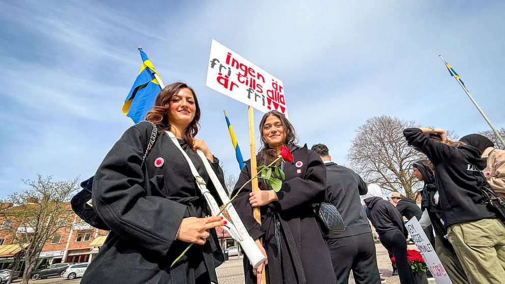Två kvinnor i svarta kappor bär skylt och rosor vid första maj-demonstration, svenska flaggor i bakgrunden.