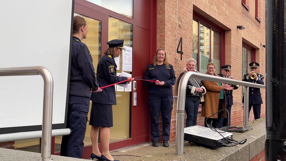 Personer i uniform står vid entrén till en byggnad under en invigning där en kvinna klipper av ett rött band. I bakgrunden finns en skärm och flera personer som applauderar, vilket indikerar att de firar öppnandet av en nyhet eller ett projekt.