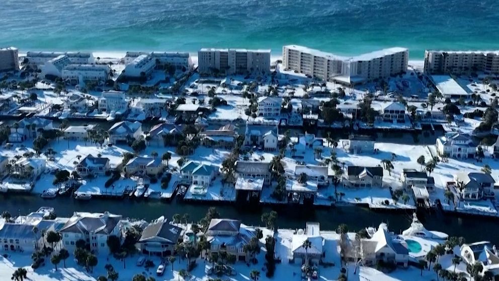 Fågelperspektiv över ett kustområde täckt av snö, där villor och bostadshus i olika storlekar syns. I bakgrunden sträcker sig det blå havet ut mot horisonten, vilket kontrasterar mot den snövita stranden och de snötäckta tak som pryder området.