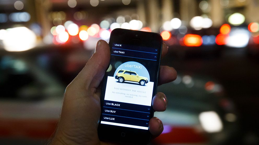 Hand håller smartphone med taxi-app, suddig stadsmiljö i bakgrunden.