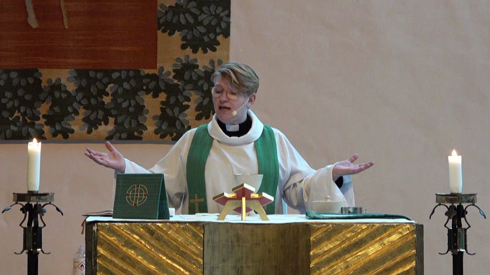 En präst i liturgiska kläder står vid ett altare med uppsträckta armar, flankerad av två tända ljus. På altaret finns en bibel eller religiös bok och ett krucifix eller en religiös symbol. Bakgrunden pryds av dekorativa element.