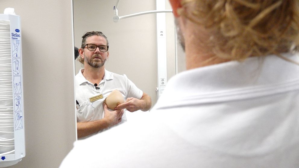 En man i vit skjorta står framför en spegel och håller en prototyp av en bröstprotes. I bakgrunden syns delar av ett rum, inklusive medicinska instrument. Mannen ser ut att demonstrera eller förklara något relaterat till protesen.