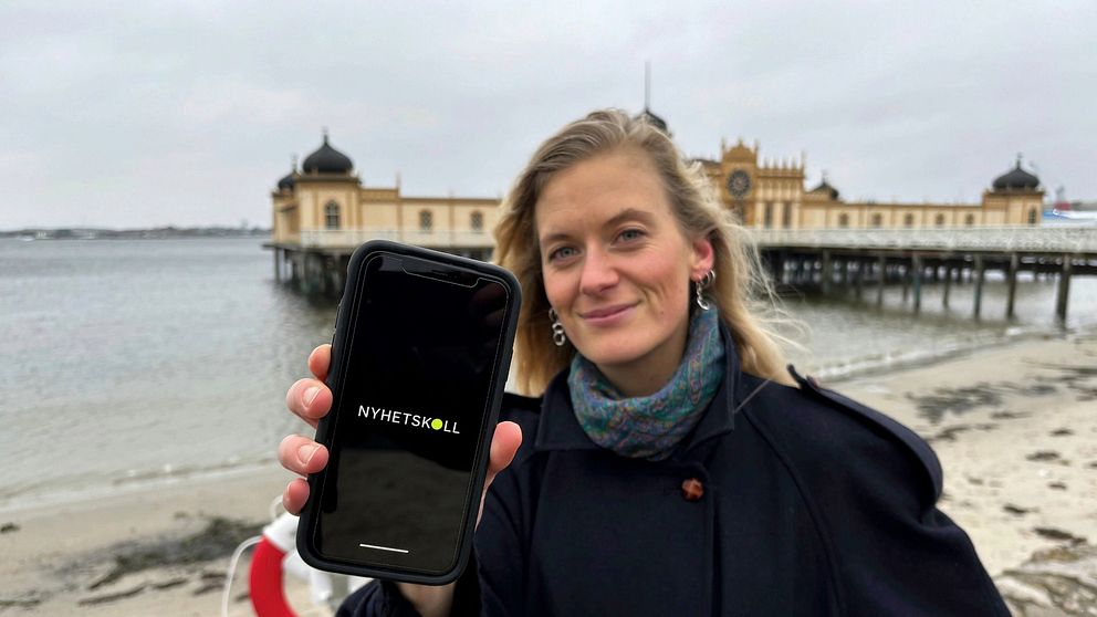 En person står vid en kust med en historisk badhusbyggnad i bakgrunden och håller upp en mobiltelefon som visar appen ”Nyhetsskolan” på skärmen.